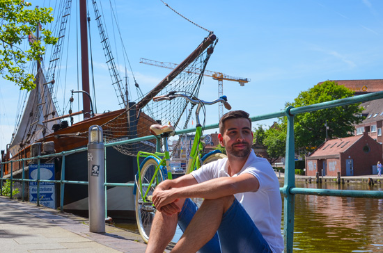 Ein junger Mann und sein Rad lehnen an einem Geländer eines Flusses, im Hintergrund ist ein Schiff.