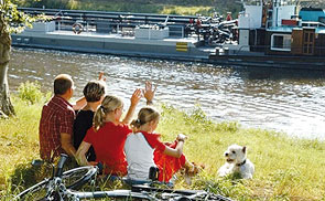 Eine Familie mit Hund sitzt auf einer Wiese an einem Fluss und winkt einem vorbeifahrendem Schiff zu.